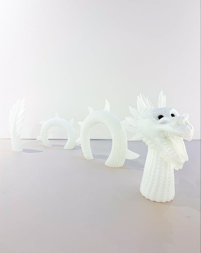 A white glass blown articulated dragon figurine, shown in a multi-segmented form with the head and neck fully visible and the body segments receding into the background.
